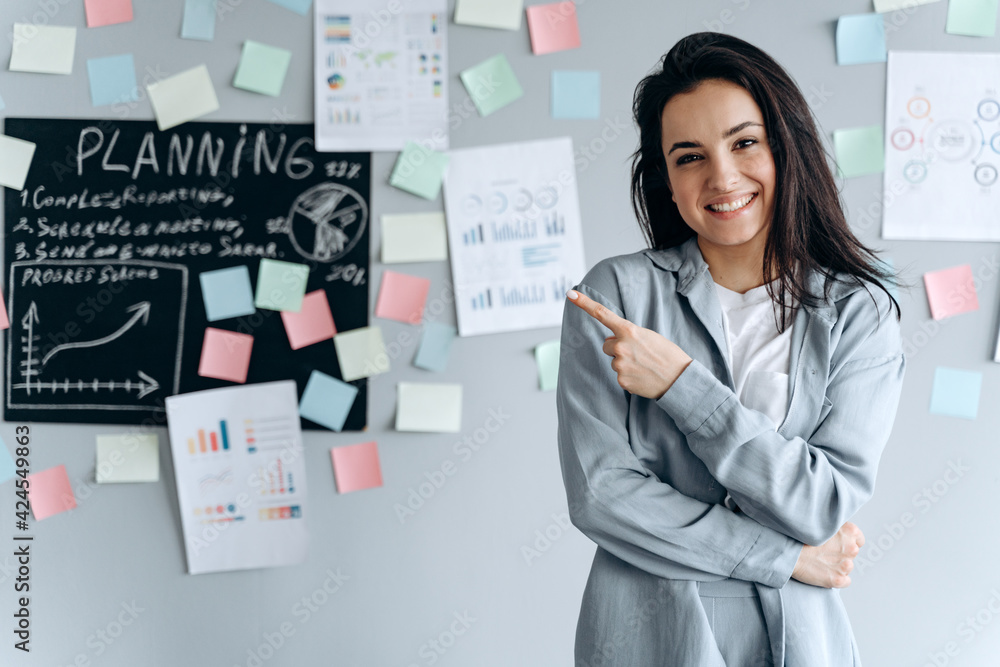 Smiling young girl in the office on a background of a gray wall with stickers. She points to something with her index finger. Smiling young girl in the office on a background of a gray wall with stickers. She points to something with her index finger.