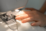 A girl with graceful hands sews a light-colored fabric on a sewing machine. Women's hands sew fabric products. Notes, a needle, seamstress. Selective focus, close-up