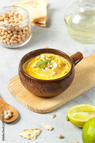 Hummus in a brown ceramic bowl on a gray table with butter in a jug, chickpeas and lime. Close-up, vertical frame