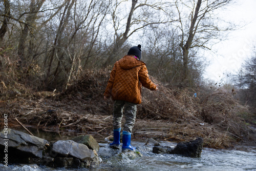 The girl in rubber boots in a brown jacket crosses the river ford, in early spring the trees around without leaves. Fun childhood © Mariyka LnT