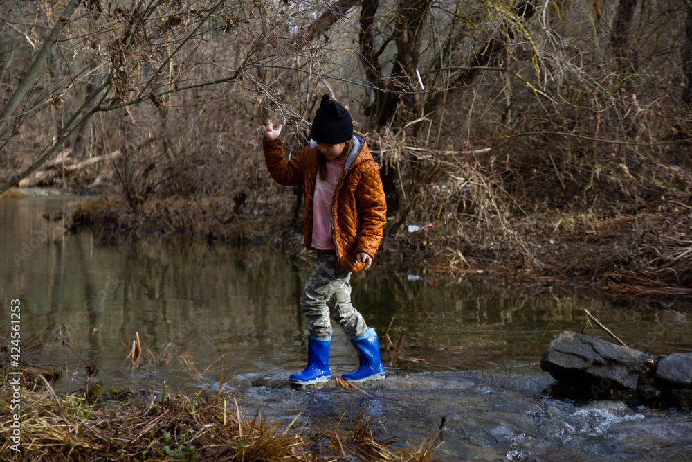 Obraz premium The girl in rubber boots in a brown jacket crosses the river ford, in early spring the trees around without leaves. Fun childhood