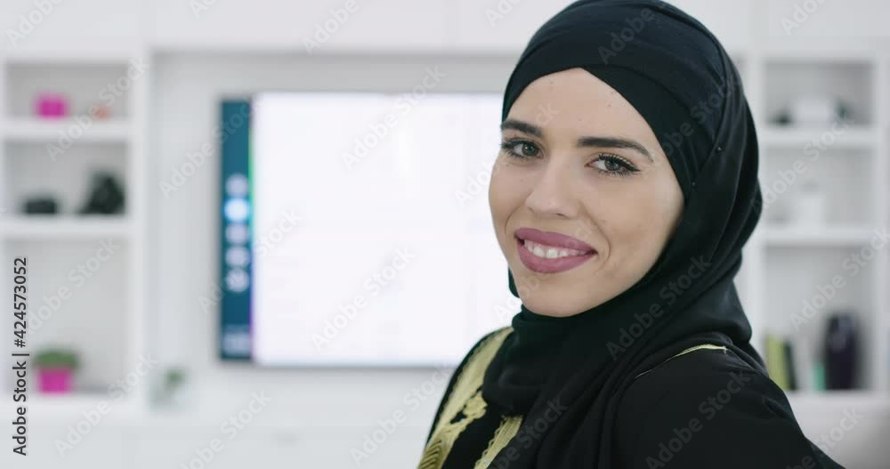 Muslim woman wearing islamic hijab clothes sitting on sofa watching TV at jhome Muslim woman wearing islamic hijab clothes sitting on sofa watching TV at jhome