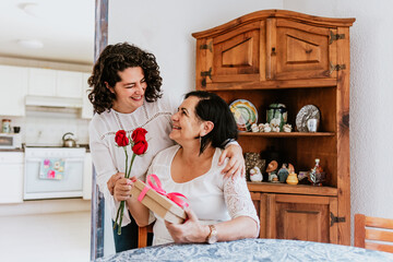 latin young woman and her mother middle age with flowers and gift box at home celebrating Happy mother's day in Mexico city