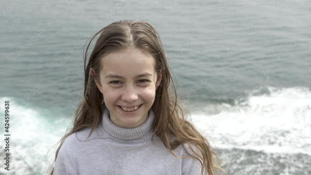 Back view. A girl with long flowing hair looks at the camera and smiles. A child smiles at the camera against the backdrop of a raging ocean. Back view. A girl with long flowing hair looks at the camera and smiles. A child smiles at the camera against the backdrop of a raging ocean.