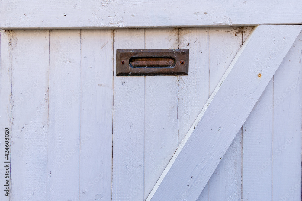 A vintage exterior wooden white door with wood panels. There's a small brass post letter or mail slot at the top and center of the wood weather shutter door. The material is worn with specs of rust.