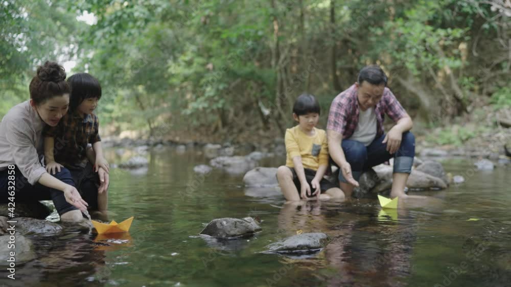An Asian family, a father and a mother teach their two sons kids how to craft a paper boat. Float in a natural forest stream. On vacation. Children's learning concept. An Asian family, a father and a mother teach their two sons kids how to craft a paper boat. Float in a natural forest stream. On vacation. Children's learning concept.