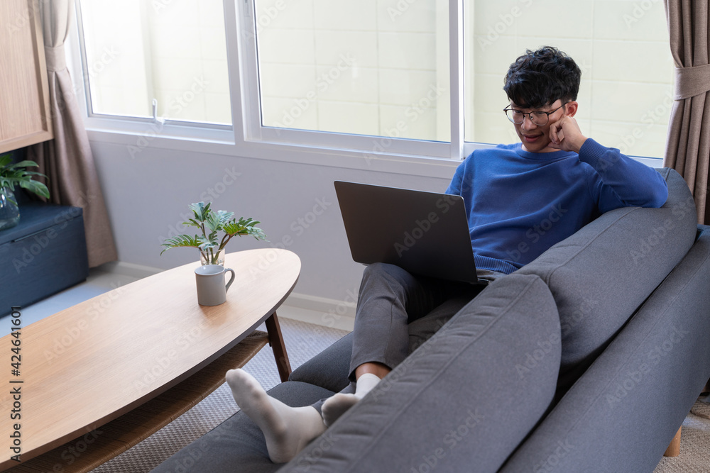 Young asian male tech user relaxing holding laptop computer and looking at the screen in living room, Remote Job or work from home concept. Young asian male tech user relaxing holding laptop computer and looking at the screen in living room, Remote Job or work from home concept.