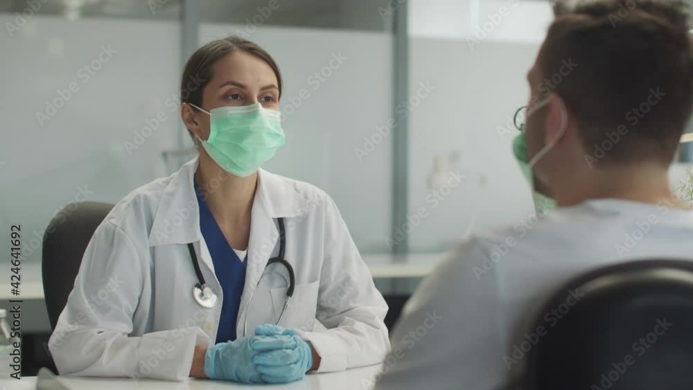 A young female doctor in a mask and gloves is receiving a patient in a medical office. Tells the young man about the need for vaccination against the virus.