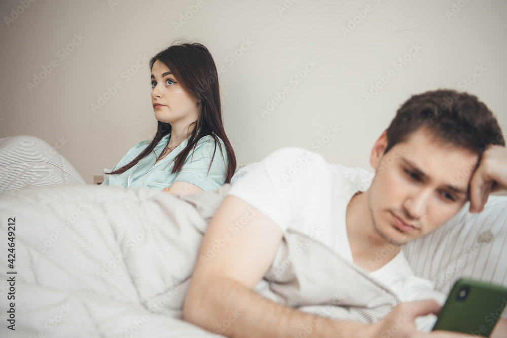 Upset caucasian woman is sad because her husband is looking in mobile while lying in bed Upset caucasian woman is sad because her husband is looking in mobile while lying in bed