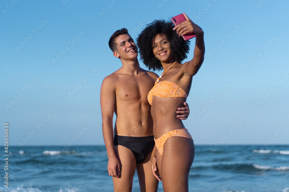 Multiracial couple in swimsuit of young people with Caucasian man and Afro-American woman taking selfie with the smartphone on the beach with the sea on the background.