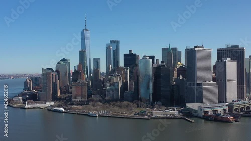 An aerial view of New York Harbor on a sunny day with blue skies. The drone camera facing lower Manhattan truck left slowly over the waters with a Staten Island Ferry docked.