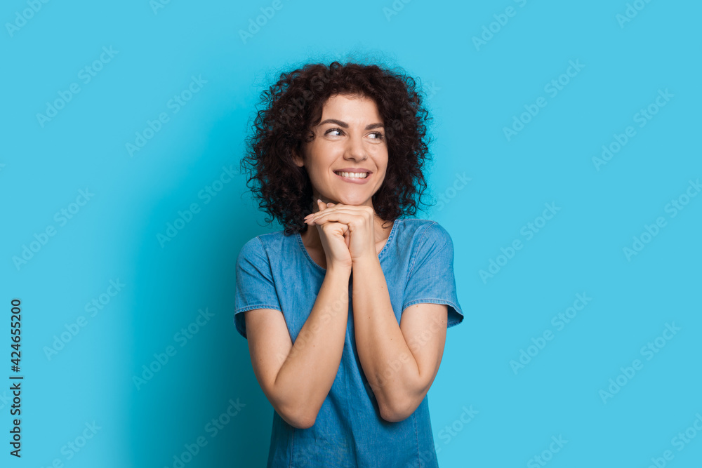 Thoughtful caucasian curly haired woman in a blue dress is dreaming about something on a studio wall looking away Thoughtful caucasian curly haired woman in a blue dress is dreaming about something on a studio wall looking away