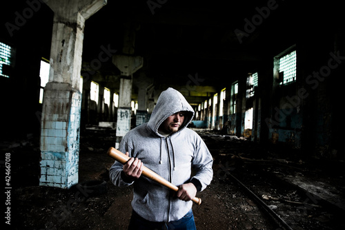 Aggressive man with a baseball bat wear hoody with hood stand inside industrial space room against window frame and beam in perspective photo