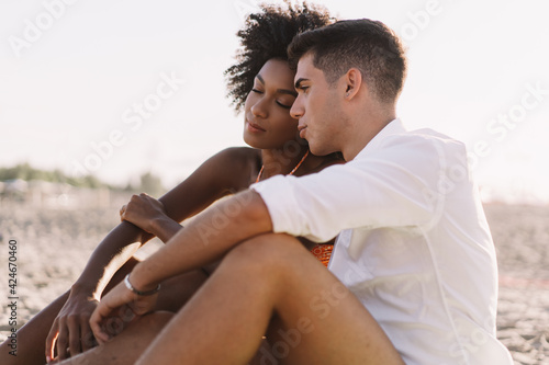 Multiracial couple of young people tenderly talking sitting on the beach at sunset. Lifestyle concept of couples in love between people of different races