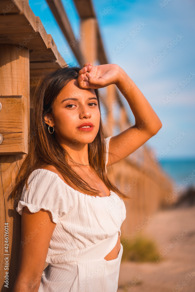 Fototapeta premium Lifestyle of a young Caucasian brunette enjoying the beach vacation in a white dress in summer, next to a wooden walkway