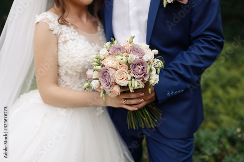 The bride and groom in wedding suits are holding together a wedding bouquet of white and lilac roses. Men's blue classic suit for men and girls white dress. Newlyweds without faces.