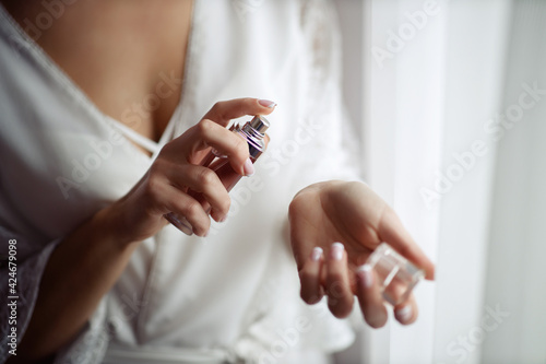 A girl in a white satin dressing gown with a neckline and a manicure french holds a bottle with perfume and splashes on her wrist