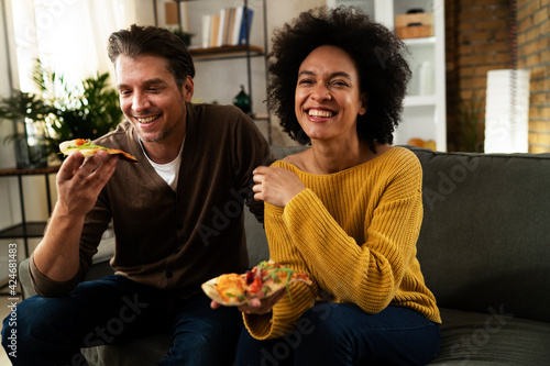 Cheerful young couple sitting on sofa at home. Happy woman and man eating pizza while watching a movie.