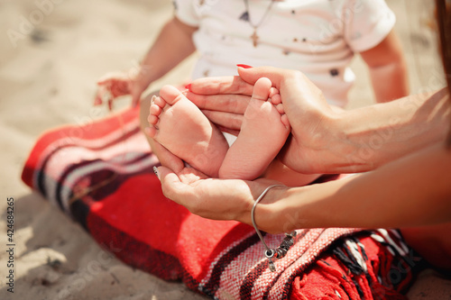 A young mother with manicure holds her little feet in her hands with her hands and shows her feet on the beach in the sand on a plaid. Close-up without a face, picnic by the sea, vacation with family