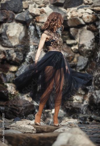 A girl with long slim legs in a shiny translucent black dress with a skirt and high-heeled shoes is dancing against the background of a waterfall and a stone wall.