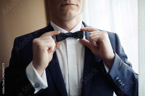 A smiling man in a white shirt and blue jacket holds a stylish black bow-tie with both hands. Groom getting dressed for a wedding in the morning