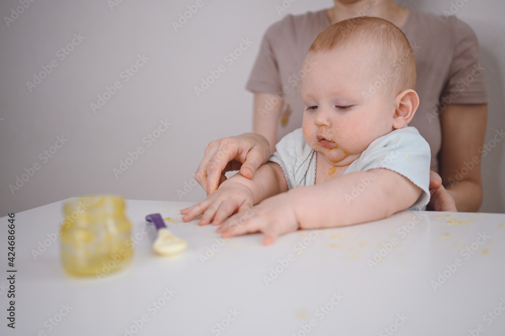 Little newborn funny baby boy learning to eat vegetable or fruit puree from glass jar with spoon. Young mother helping little son eat first food Little newborn funny baby boy learning to eat vegetable or fruit puree from glass jar with spoon. Young mother helping little son eat first food