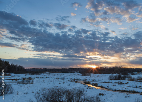 Bright rays of the sun on the horizon. Sunset  the sun shines through the clouds. Snow and bushes in the foreground. Early spring. Sunlight is reflected in the river.