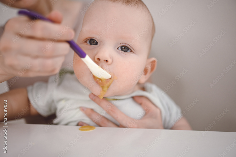 Little newborn funny baby boy learning to eat vegetable or fruit puree from glass jar with spoon. Young mother helping little son eat first food Little newborn funny baby boy learning to eat vegetable or fruit puree from glass jar with spoon. Young mother helping little son eat first food