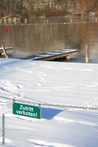 Sign on a old chain saying: No entry ( german: zutritt verboten ). White letters on green board. Frozen lake with pier in the background. Winter in Germany, Max-eyth-see.