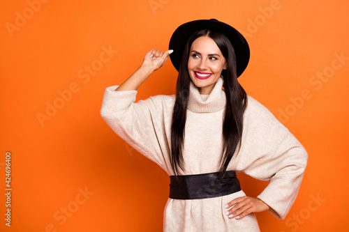 Photo of shiny charming young woman wear beige sweater arm headwear looking empty space isolated orange color background