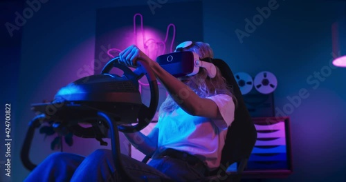 A girl is playing racing game in VR. She is sitting on the gaming chair with a steering wheel and pedals. A girl wears VR headset. Chair is located in the living room with red and blue neon lighting.
