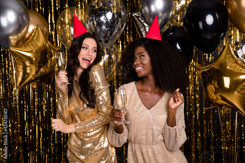 Photo of two optimistic brunette curly hairdo ladies with balloons glass wear hat nice dress isolated on gold background