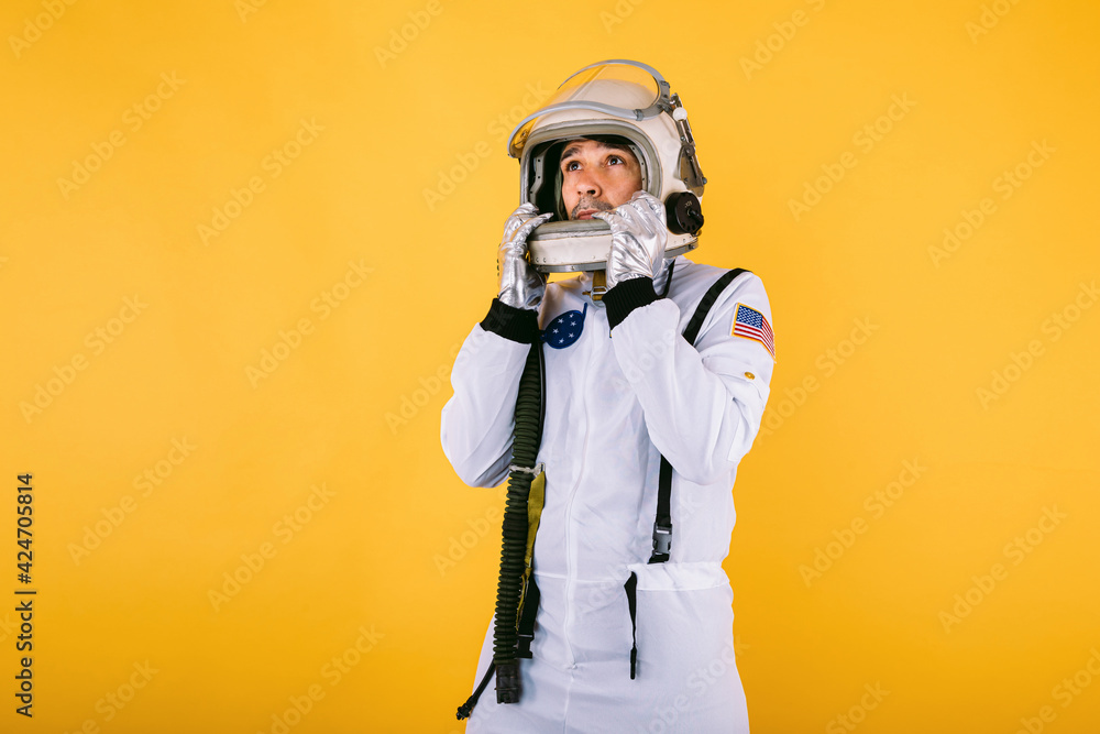 Male cosmonaut in spacesuit and helmet, holding helmet with hands and looking to the left, on yellow background. Male cosmonaut in spacesuit and helmet, holding helmet with hands and looking to the left, on yellow background.