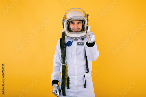 Male cosmonaut in spacesuit and helmet, raising thumb, on yellow background. © Davidbenito