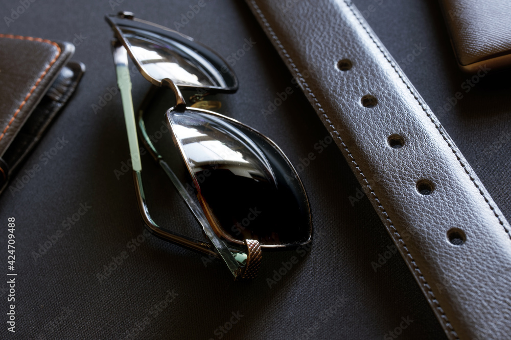 A black leather belt, sunglasses, a note book and a wallet lie on a dark background. Men's accessories and masculinity A black leather belt, sunglasses, a note book and a wallet lie on a dark background. Men's accessories and masculinity