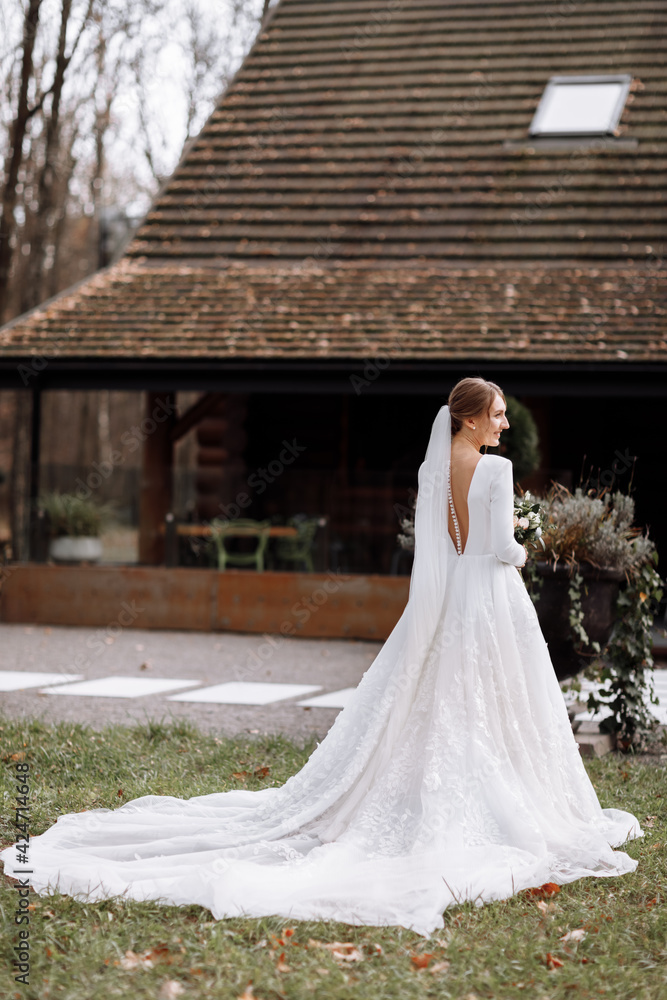 Obraz premium bride in a long white dress is standing in the park and holding a wedding bouquet of roses flowers and greens. young woman with hairstyle, makeup and long veil outdoors near hotel