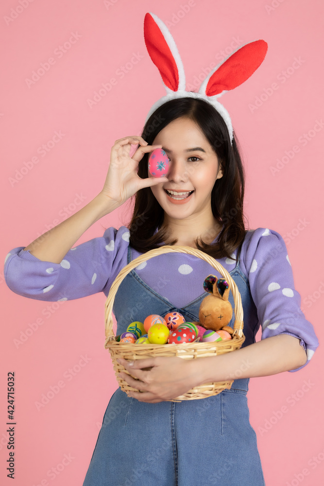 Young Asian woman wearing a Bunny hairband, holding an Easter egg, covering her eyes and hold a basket full of colorful eggs with a smiling face. The concept easter eggs festival with pink background Young Asian woman wearing a Bunny hairband, holding an Easter egg, covering her eyes and hold a basket full of colorful eggs with a smiling face. The concept easter eggs festival with pink background