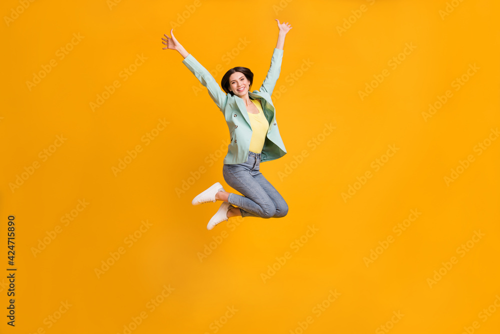Full length photo of adorable sweet young lady dressed turquoise jacket jumping rising arms isolated yellow color background