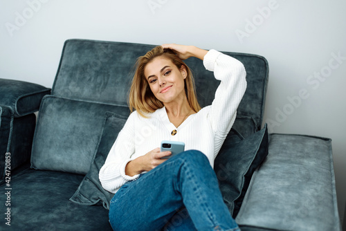 Happy young woman holding cellphone sitting on sofa and looking at camera. Female customer using tech apps on smartphone at home, enjoying easy online shopping © etonastenka