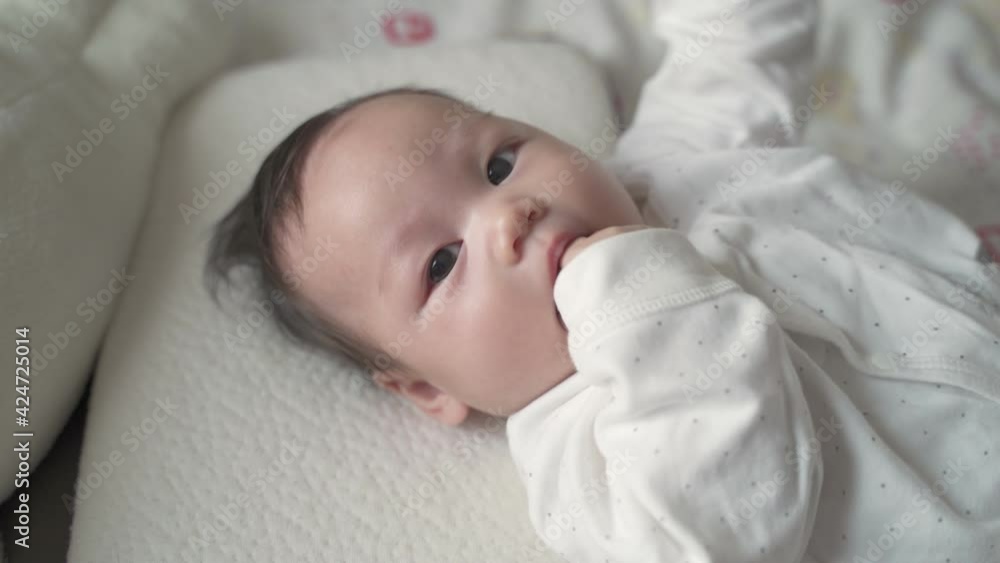 asian baby infant laying down on white soft bed sucking hand and fingers. 3 months old baby facial expression. sign and symptom of hungry in kid. asian baby infant laying down on white soft bed sucking hand and fingers. 3 months old baby facial expression. sign and symptom of hungry in kid.