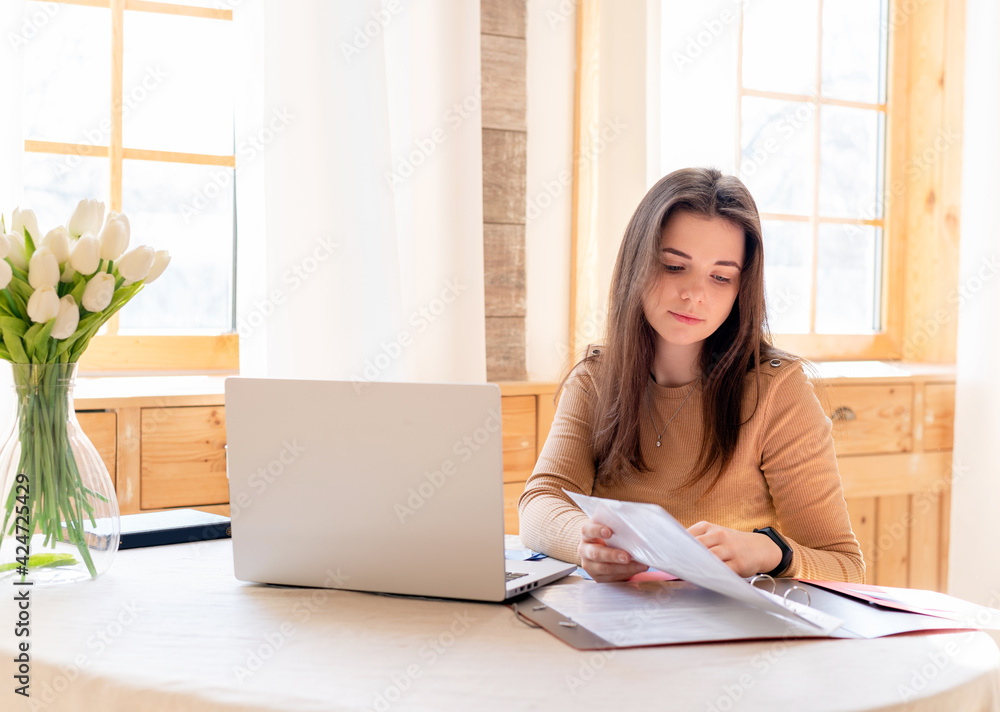 Teen girl with laptop doing homework in bright kitchen with flowers on the table. education and business concept. remote work and freelance work. student.