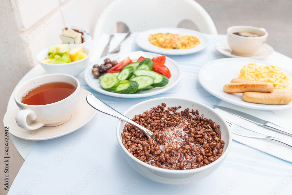 A rich breakfast is served in the hotel restaurant on the outdoor terrace. Chocolate cornflakes with milk and vegetables and tea are on the table.