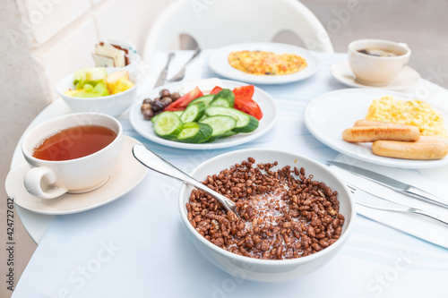 A rich breakfast is served in the hotel restaurant on the outdoor terrace. Chocolate cornflakes with milk and vegetables and tea are on the table.