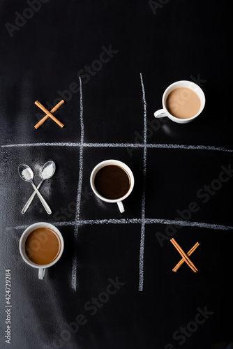 top view of tic tac toe game with spoons, cinnamon sticks and cups of coffee on black