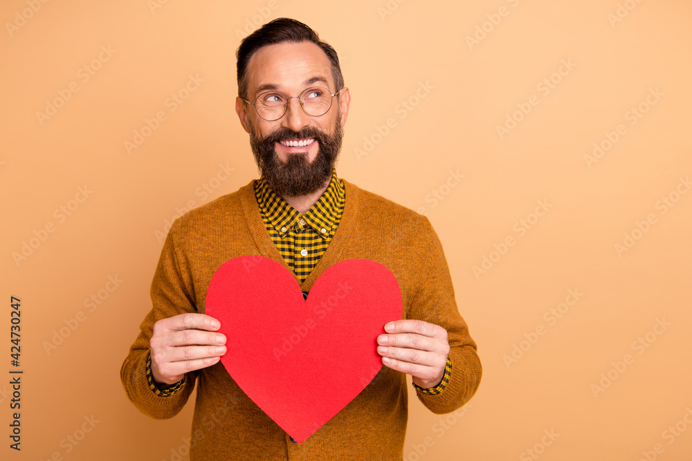 Photo of romantic man hold red heart figure look empty space wear specs brown cardigan isolated beige color background Photo of romantic man hold red heart figure look empty space wear specs brown cardigan isolated beige color background