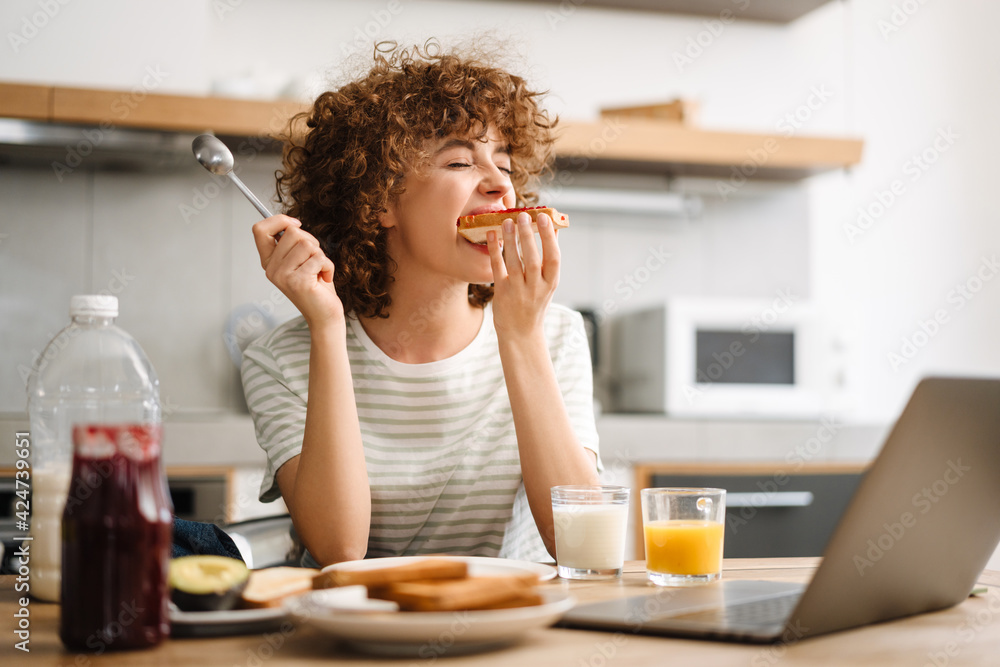 Smiling young woman using laptop while having breakfast at home kitchen - obrazy, fototapety, plakaty 