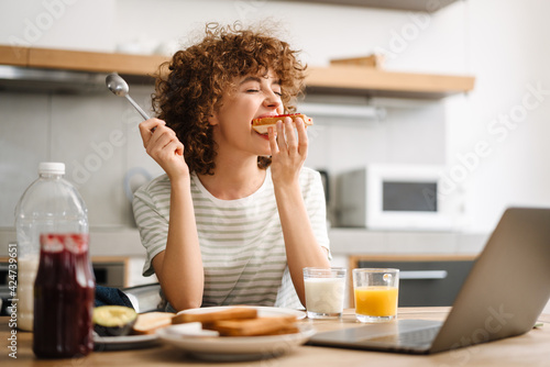 Print op canvas Smiling young woman using laptop while having breakfast at home kitchen