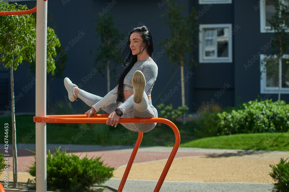 Fototapeta premium A young woman goes in for sports on the sports ground, sits on the uneven bars in a twine. Stretching, sport, workout outdoor concept