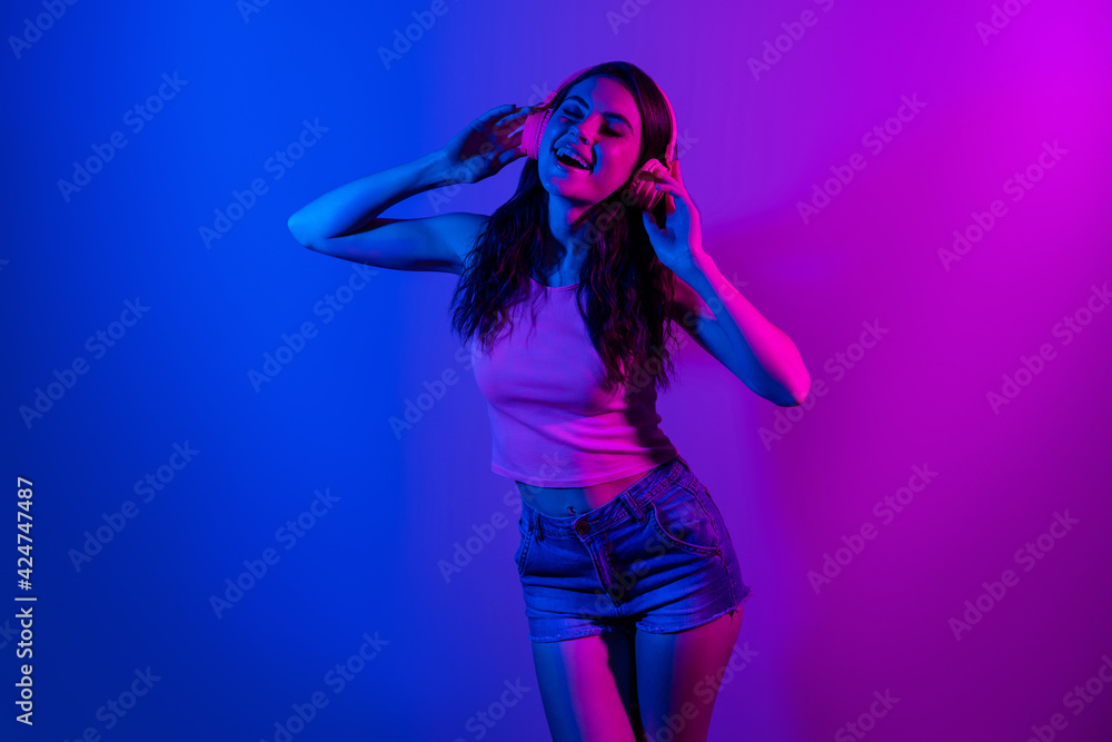 Photo of young funky beautiful smiling woman in headphones dancing isolated on glowing blue color background Photo of young funky beautiful smiling woman in headphones dancing isolated on glowing blue color background