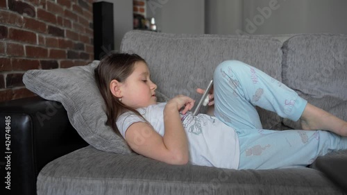 A little girl lies at home on the couch and looks into a smartphone. The child plays and watches the video. The girl has bare feet. The child is wearing pajamas. 4K